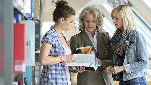 Two Female Students with their Professor Discussing a Book in Library