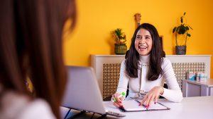 A professional businesswoman in a white outfit is actively discussing ideas during a meeting in a modern office with a vibrant yellow wall and stylish decor. She gestures expressively while holding a pen, emphasizing her points. A notebook and a laptop are visible on the table, highlighting a collaborative and dynamic workspace. The setting includes plants and a guitar in the background, adding a creative and inviting atmosphere.