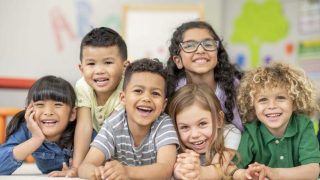 A small group of 6 Kindergarten students lay stacked on top of one another, on their classroom floor, as they pose for a portrait. They are each dressed casually and are smiling for the photo.